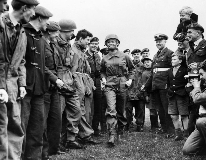 French actress Brigitte Bardot, in parachutist's equipment, walks among Royal Air Force boys, who have cancelled their leave, during the shooting of "Babette goes to war"