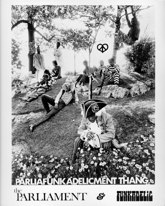 George Clinton (rear left), Fuzzy Haskins (far back below "Peace & Love" symbol), Tiki Fulwood (laying down on grass front left), and Grady Thomas (laying down second from left) of the funk band Parliament-Funkadelic pose for a portrait
