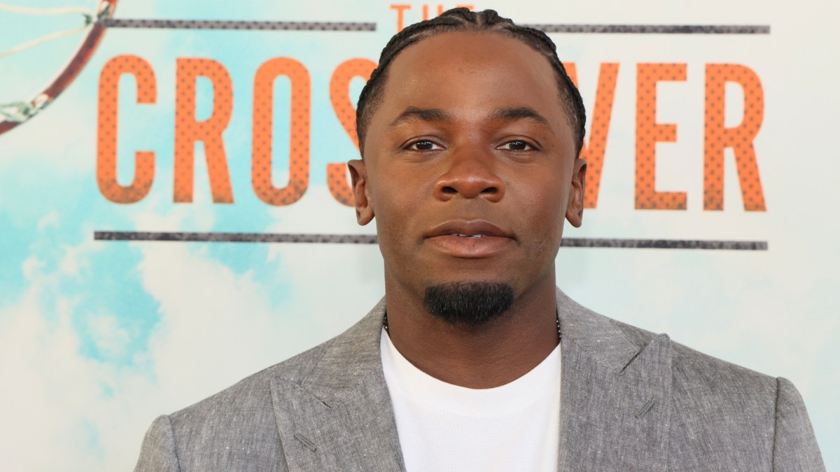 Derek Luke wears a white shirt and grey blazer.
