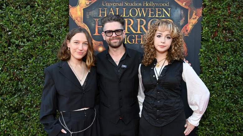 Jack Osbourne stands with two of his children in front of a green wall.